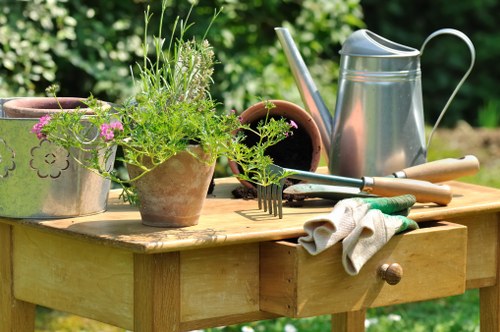 A wooden garden table in an outdoor setting holds various gardening tools and planters, including a large metal watering can, a small clay pot filled with green herbs or plants, and a terracotta flowerpot. The table surface displays a pair of gardening gloves and a hand fork, suggesting recent gardening activity. In the background, blurred lush green foliage indicates a healthy garden environment, likely in a residential outdoor space within Docklands. The scene is well-lit by natural daylight, highlighting the textures of the wooden table, metal watering can, and vibrant green plants, emphasizing the practical and organized aspect of outdoor garden maintenance typical of local garden care services. The setting shows a tidy arrangement for gardening tasks, embodying a well-maintained garden area, arranged for planting, watering, and general outdoor care, suitable for gardening and landscaping services offered in the Docklands area.