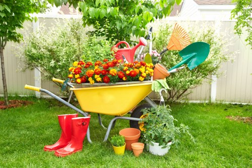 The image depicts a vibrant garden scene with a yellow wheelbarrow filled with colorful flowering plants, placed on a well-maintained green lawn. Surrounding the wheelbarrow are various gardening tools and accessories, including a red watering can, a pair of red rubber boots, and several small plant pots in terracotta and white. To the right, a potted green plant with lush foliage is visible, accompanied by a garden rake and a small gardening fork leaning against the wheelbarrow. In the background, there is a white fence with dense leafy trees and shrubs providing a lush backdrop, suggesting a private backyard or outdoor space in a residential area, possibly in London or nearby areas like Docklands. The scene is well-lit, indicating daytime with natural sunlight, and the overall setting reflects active gardening and outdoor maintenance activity suitable for a garden care service in the Docklands area.