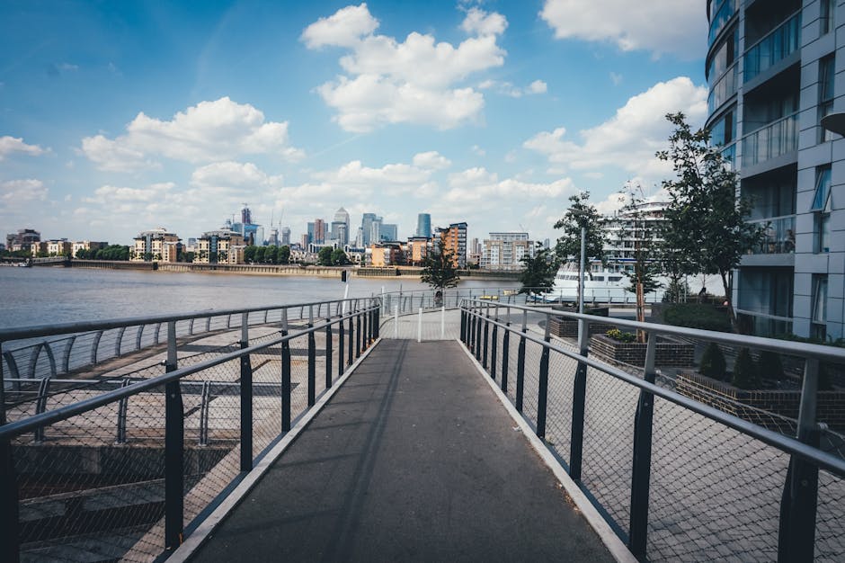A wide, paved pedestrian walkway with metal railings on both sides extending over the dockside area along the River Thames in Canary Wharf. On the right, there are modern high-rise residential buildin