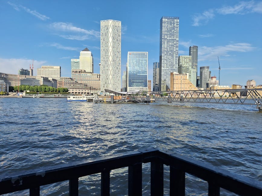 A wide, paved pedestrian walkway with metal railings on both sides extending over the dockside area along the River Thames in Canary Wharf. On the right, there are modern high-rise residential buildin